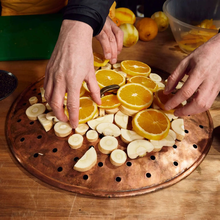 Two hands arrange slices of orange and parsnip on a round tray, perhaps prepping an infusion with James Gin Navy Strength 700mL 114 Proof by James Gin, while more fresh produce is scattered across the wooden table in the background.
