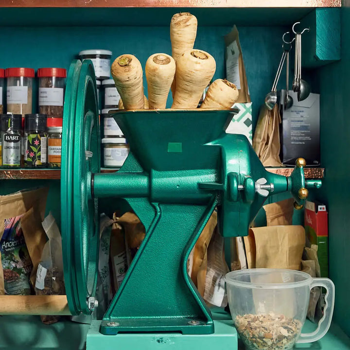 A green metal hand-crank grinder packed with whole parsnips sits beside spice jars and measuring spoons—ideal for creating James Gin Asian Parsnip 700mL 80 Proof by James Gin, which comes with a souvenir gift key chain.