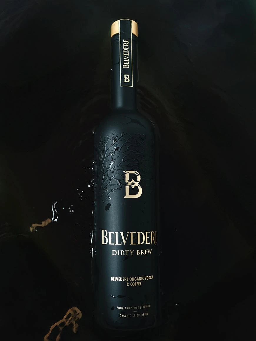 A matte black bottle of Belvedere Dirty Brew Organic Coffee Flavored Vodka by Belvedere Vodka, accented with gold lettering, rests on a dark surface amid coffee splashes. The label highlights “Organic Vodka & Coffee.”.