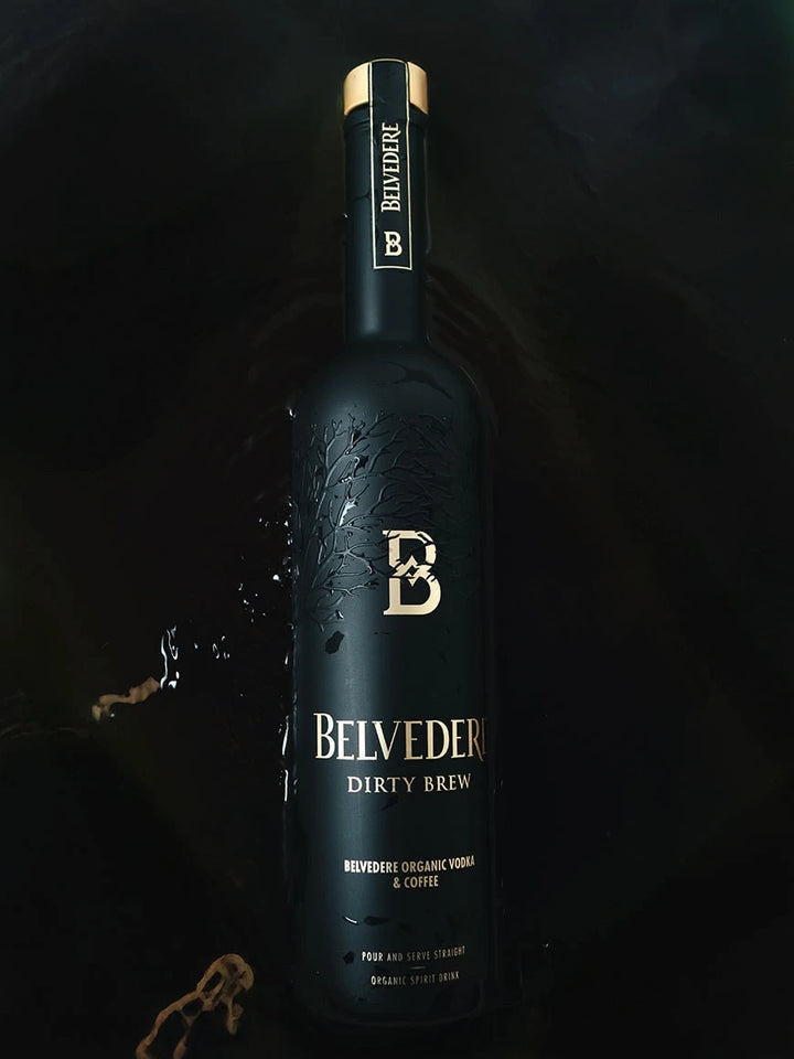 A matte black bottle of Belvedere Dirty Brew Organic Coffee Flavored Vodka by Belvedere Vodka, accented with gold lettering, rests on a dark surface amid coffee splashes. The label highlights “Organic Vodka & Coffee.”.