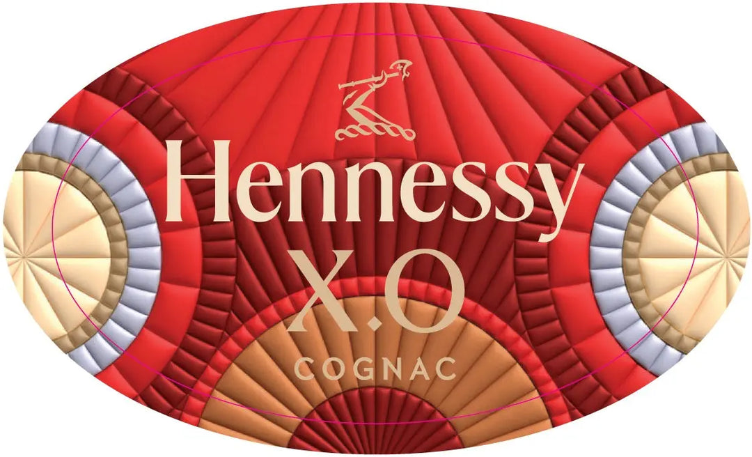 An oval label with red, gold, and beige geometric patterns bears the Hennessy X.O. Year of the Fire Lunar New Year Goat by Raxxy Cognac Limited Edition branding and an arm holding an axe logo, celebrating luxury French cognac.