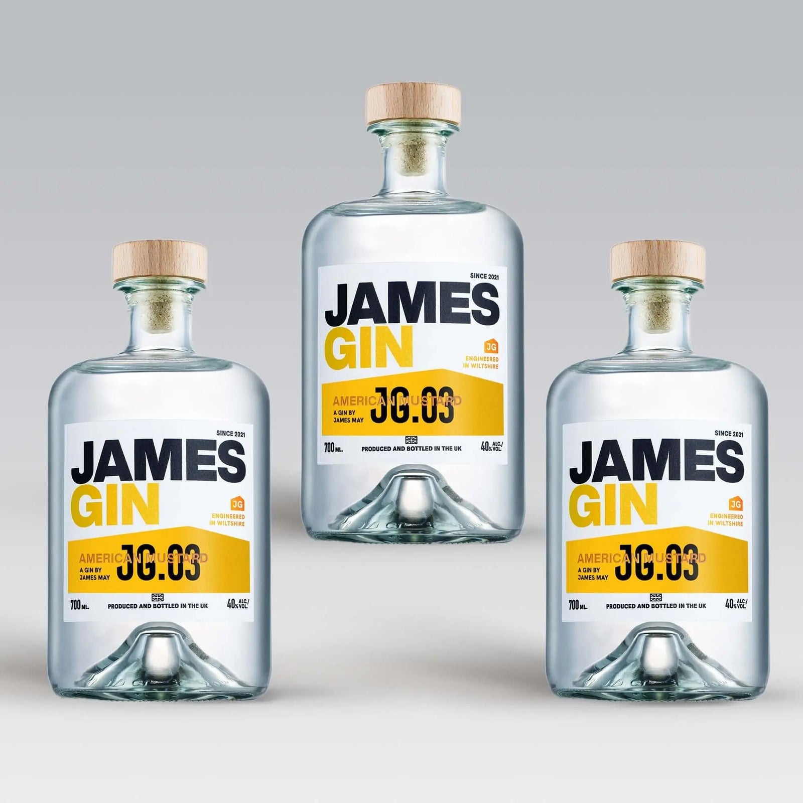 Three bottles of James Gin US "The Big, Beautiful Bundle" with wooden caps are arranged in a triangle on a light gray background. Each features a white label with bold black and yellow text, plus a souvenir gift key chain.