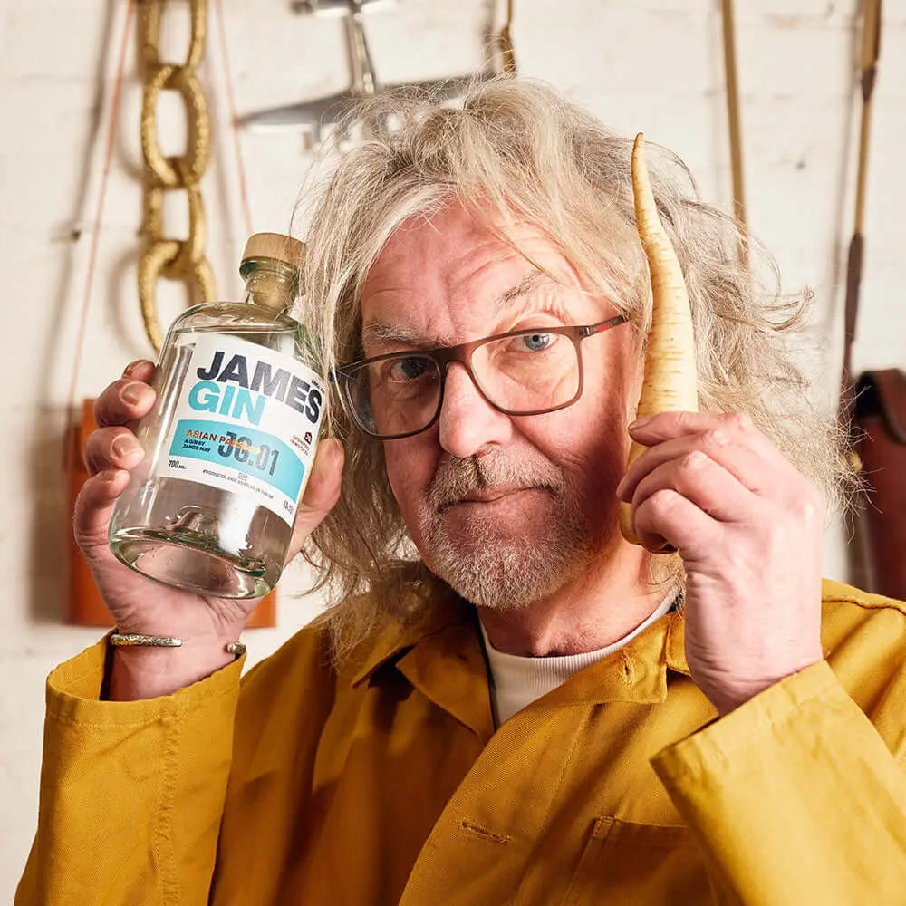 A man with gray hair and glasses, in a yellow shirt, holds a James Gin The Tupac 2 Bottle Set W/ Souvenir Gift Key Chain by James Gin US in one hand and a parsnip in the other, standing before a white wall with hanging tools.