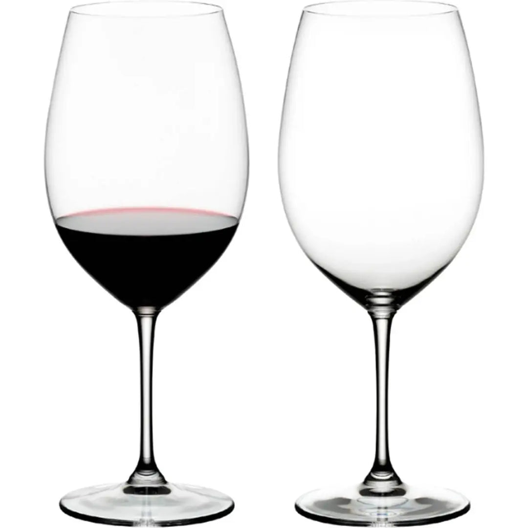 A set of two Riedel Vinum Red Wine glasses by Riedel—one filled with red wine and the other empty—are displayed side by side against a plain white background.